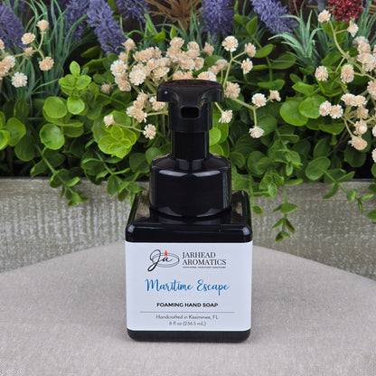 A black pump bottle of Jarhead Aromatics Maritime Escape Foaming Hand Soap rests on a beige surface, with vibrant artificial flowers and green leaves in the background.