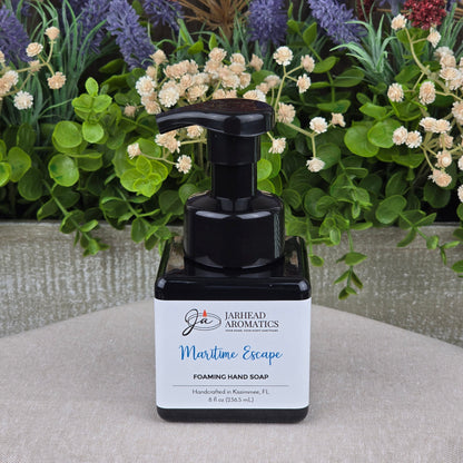 A bottle of Jarhead Aromatics Maritime Escape Foaming Hand Soap rests on a light surface, set against artificial flowers and greenery.