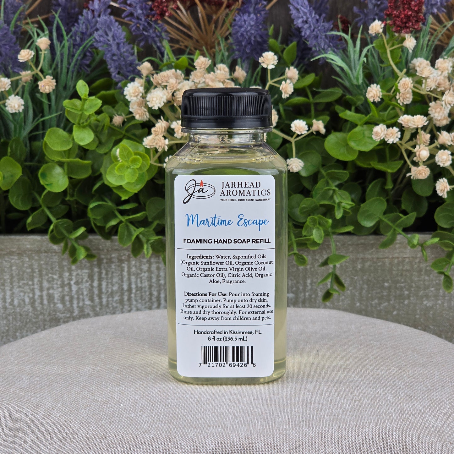 A clear bottle of Jarhead Aromatics Maritime Escape Foaming Hand Soap sits on a gray surface with a coastal scent. The label displays plant-based oils, ingredients, and directions, as green plants and purple flowers blur in the background.