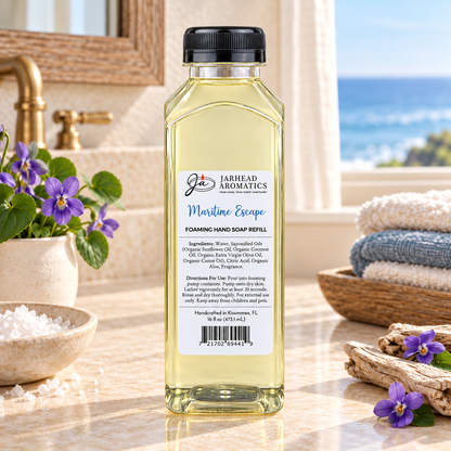 Clear bottle of Maritime Escape Handmade Foaming Hand Soap with a label featuring product ingredients, usage instructions, and a barcode, sitting on a marble countertop next to violet flowers and folded towels.