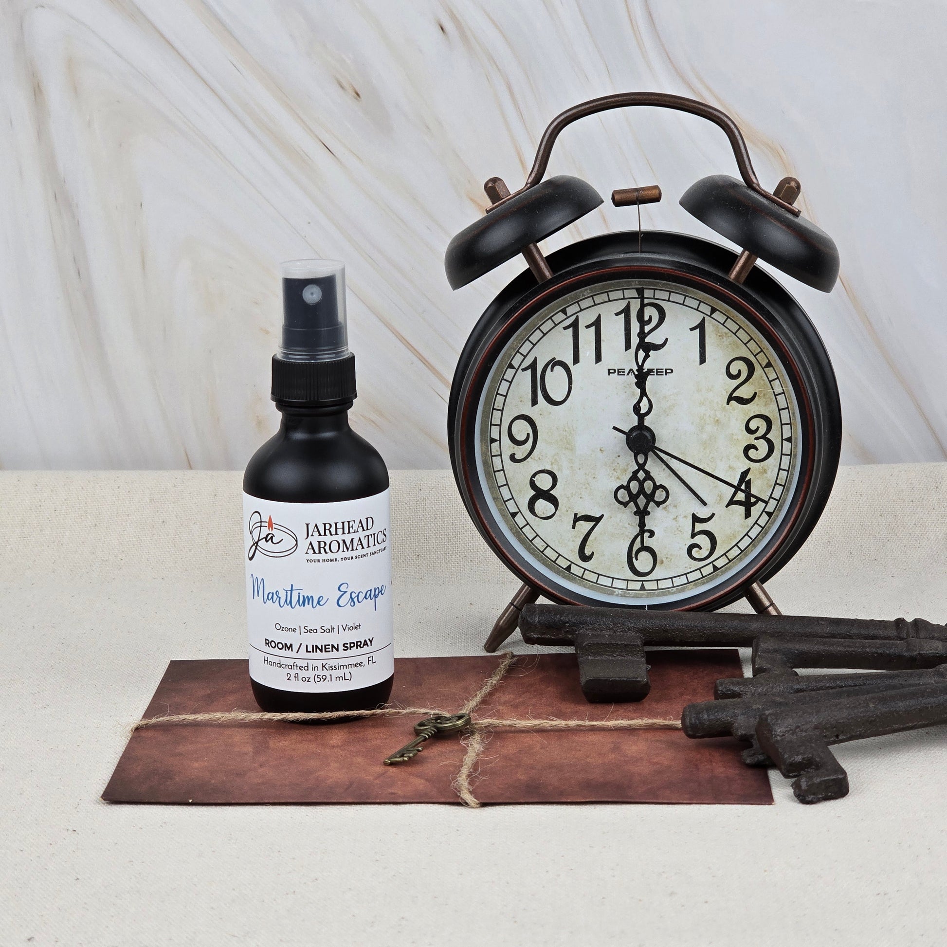 A vintage alarm clock sits beside Jarhead Aromatics’ Maritime Escape Room & Linen Spray—Sea Salt & Coastal Spa scent—along with old keys and a brown, string-tied envelope on a light surface.