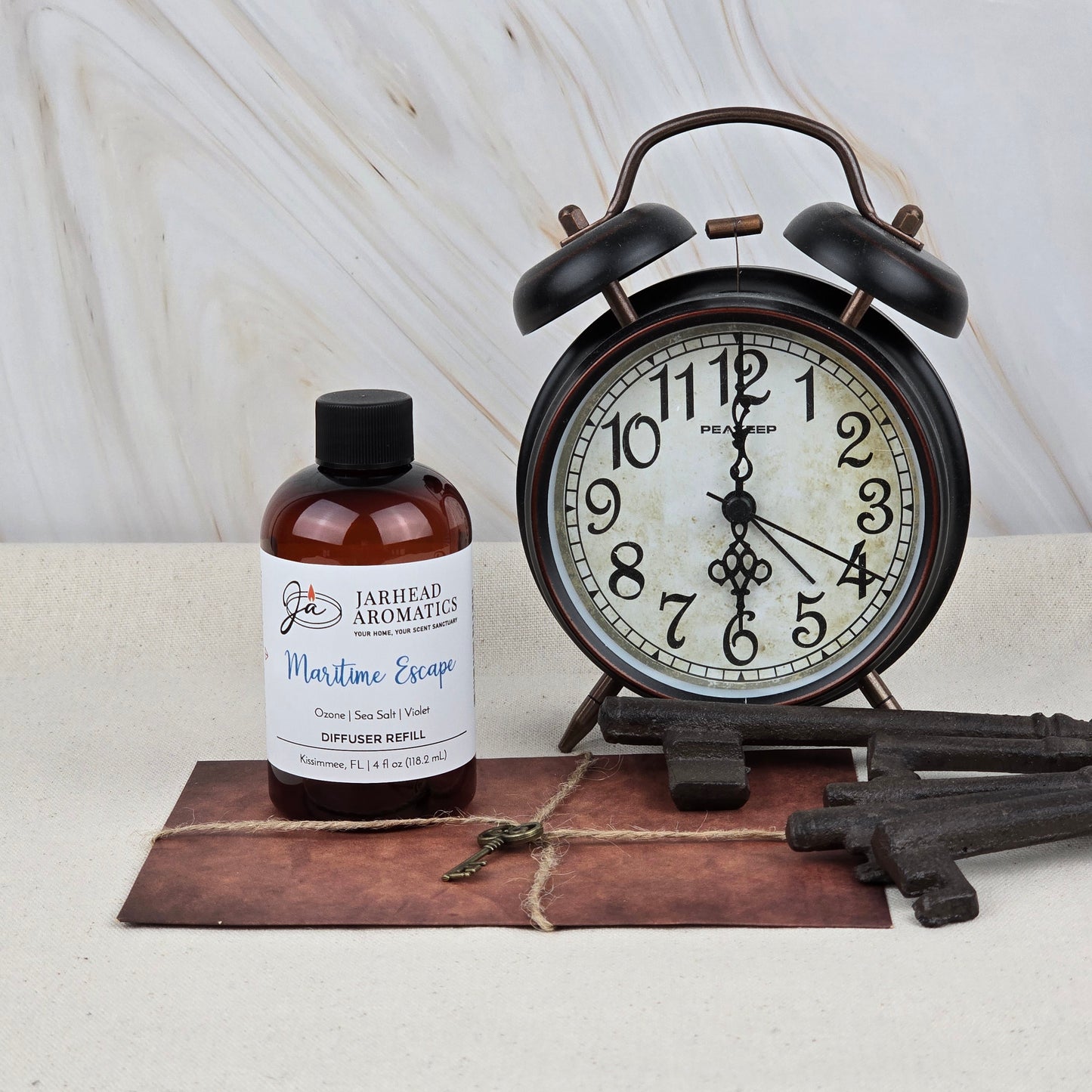 A bottle of Jarhead Aromatics Maritime Escape Diffuser Refill (Sea Salt, Ozone & Cedar) sits beside a vintage black alarm clock, three antique keys, and an old envelope with a small key on it, all on a light surface.