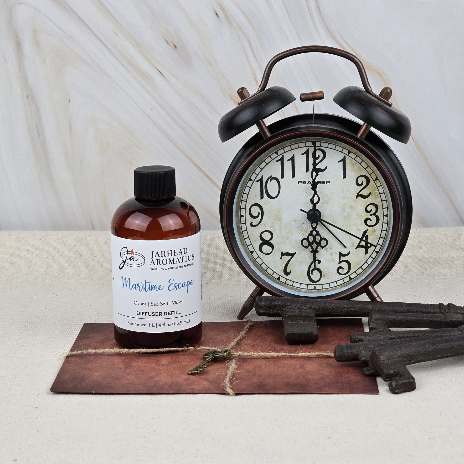 A bottle of Jarhead Aromatics Maritime Escape Diffuser Refill (Sea Salt, Ozone & Cedar) sits beside a vintage black alarm clock, three antique keys, and an old envelope with a small key on it, all on a light surface.