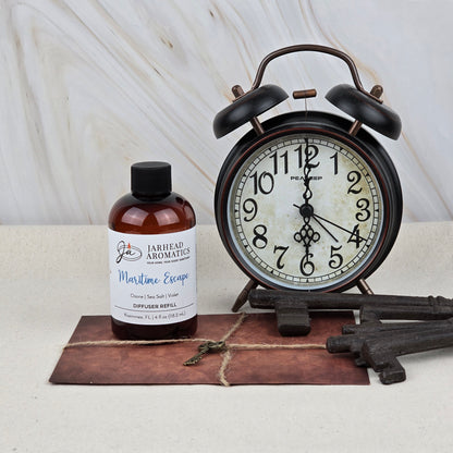 A bottle of Jarhead Aromatics Maritime Escape Diffuser Refill (Sea Salt, Ozone & Cedar) sits beside a vintage black alarm clock, three antique keys, and an old envelope with a small key on it, all on a light surface.