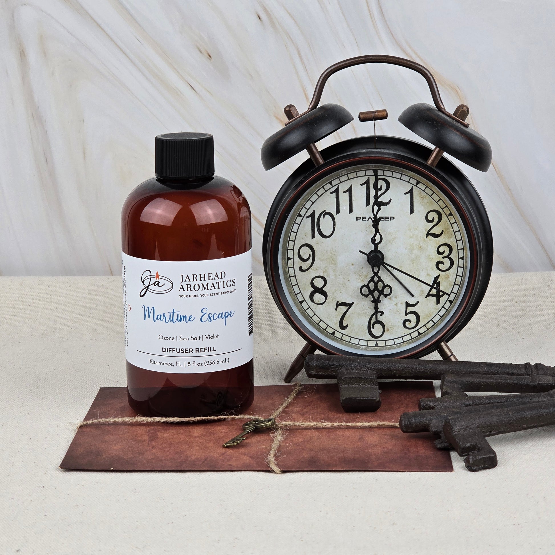 A brown bottle of Maritime Escape Diffuser Refill | Sea Salt, Ozone & Cedar by Jarhead Aromatics sits beside a black vintage alarm clock and antique keys on brown leather, evoking a coastal spa ambiance.