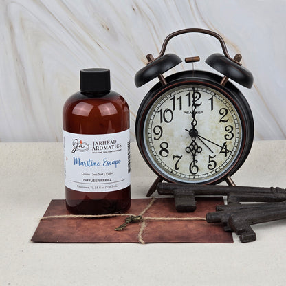 A brown bottle of Maritime Escape Diffuser Refill | Sea Salt, Ozone & Cedar by Jarhead Aromatics sits beside a black vintage alarm clock and antique keys on brown leather, evoking a coastal spa ambiance.