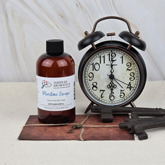 A brown bottle of Maritime Escape Diffuser Refill | Sea Salt, Ozone & Cedar by Jarhead Aromatics sits beside a black vintage alarm clock and antique keys on brown leather, evoking a coastal spa ambiance.