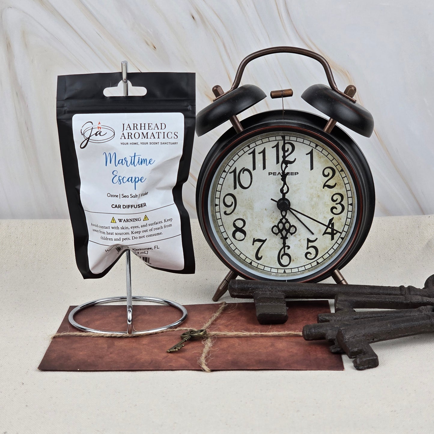 A black alarm clock, Jarhead Aromatics' Maritime Escape Car Diffuser, large antique keys, and a small key rest on a brown leather mat against a light background, evoking the clean ocean breeze and sea salt fragrance.