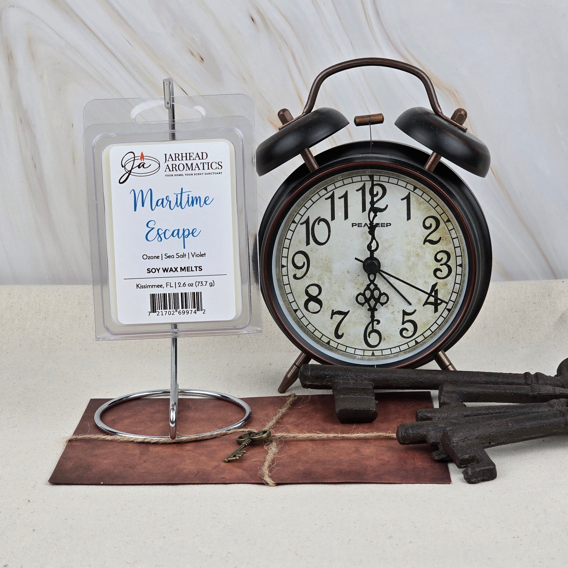 A pack of Jarhead Aromatics' Maritime Escape Soy Wax Melts, in Fresh Ocean Breeze & Sea Salt scent, is displayed on a stand next to a vintage alarm clock, two decorative keys, and an anchor charm atop a marble-like surface.
