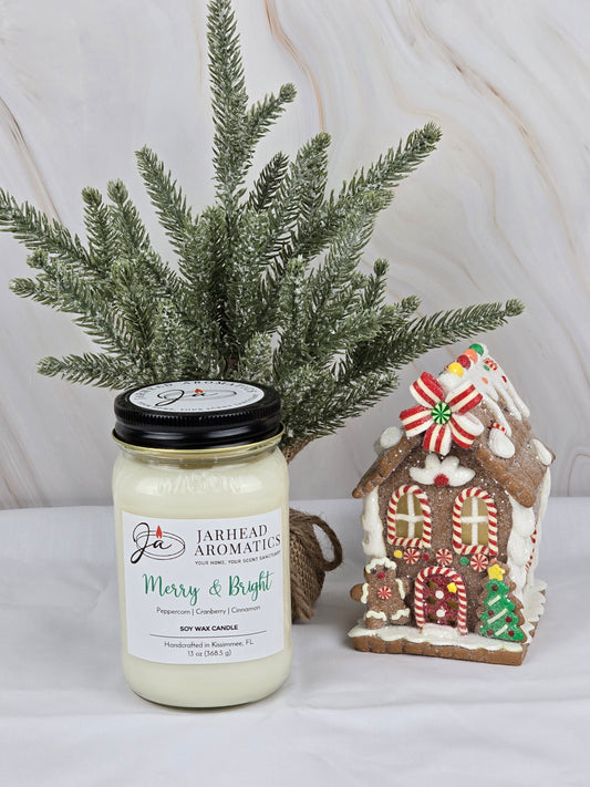 A Merry & Bright soy wax candle by Jarhead Aromatics, exuding a holiday fragrance, sits beside a gingerbread house with a small evergreen branch against a light background.