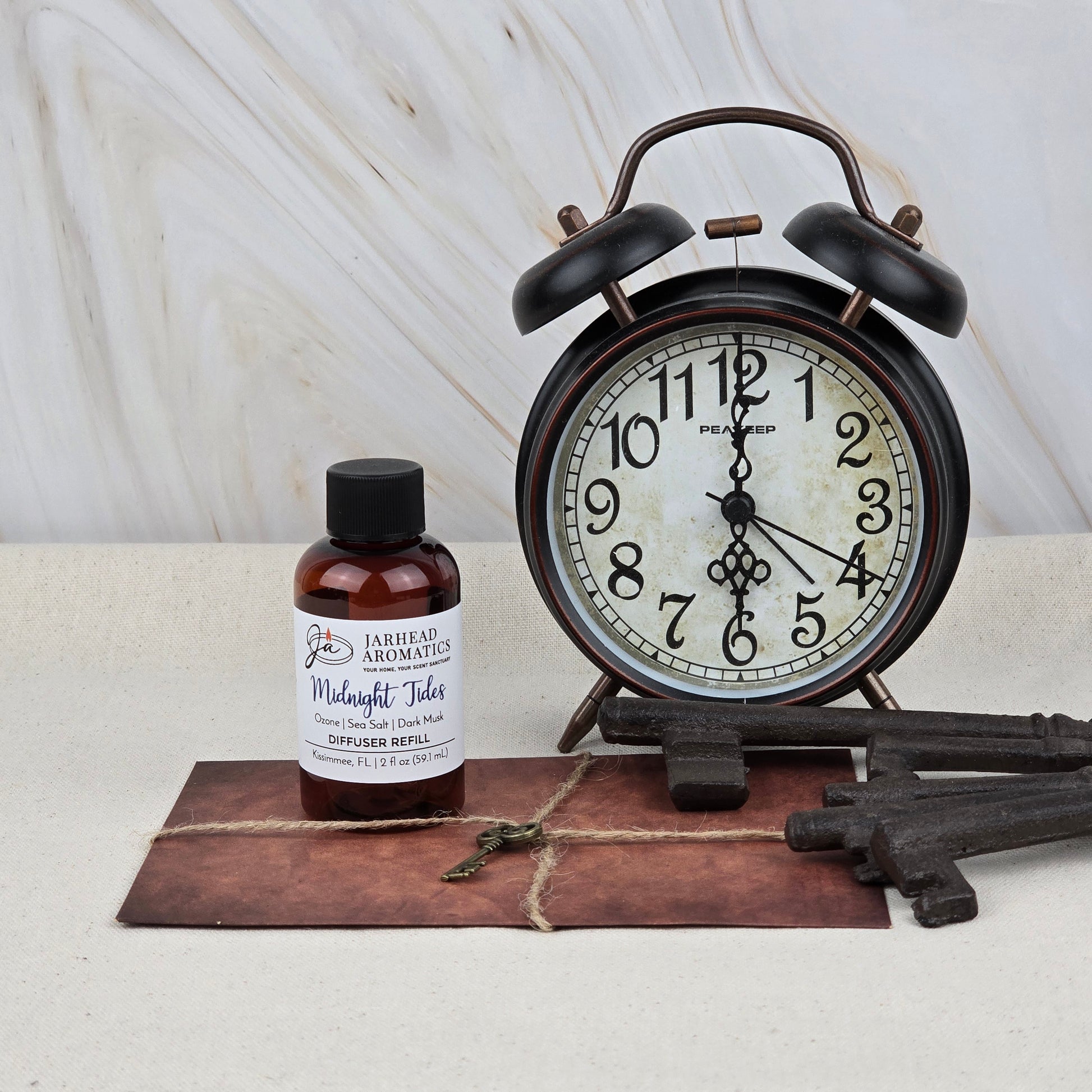 A vintage black alarm clock shows 2:36 beside a bottle of Jarhead Aromatics Midnight Tides Diffuser Refill—Sea Salt, Dark Musk & Sandalwood. Large antique keys and a brown string envelope sit on the marble table.