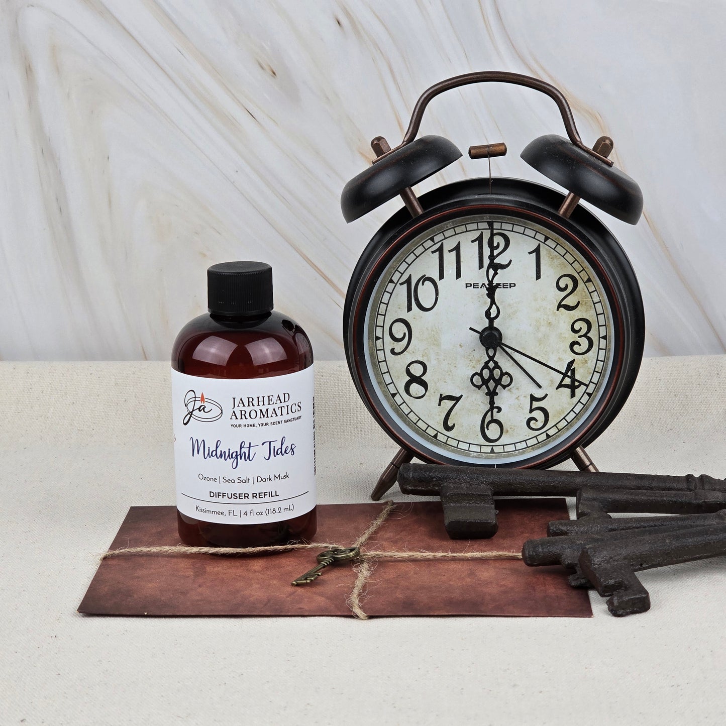 A brown bottle of Jarhead Aromatics Midnight Tides Diffuser Refill sits on a leather-bound notebook beside vintage keys and a black retro alarm clock set to 9:09, all arranged on a light surface with a marble-patterned background.