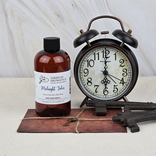 A brown bottle of Jarhead Aromatics Midnight Tides Diffuser Refill—Sea Salt, Dark Musk & Sandalwood—stands by a vintage black alarm clock at 6:09, with old keys and a brown pouch on a light surface against a marbled background.