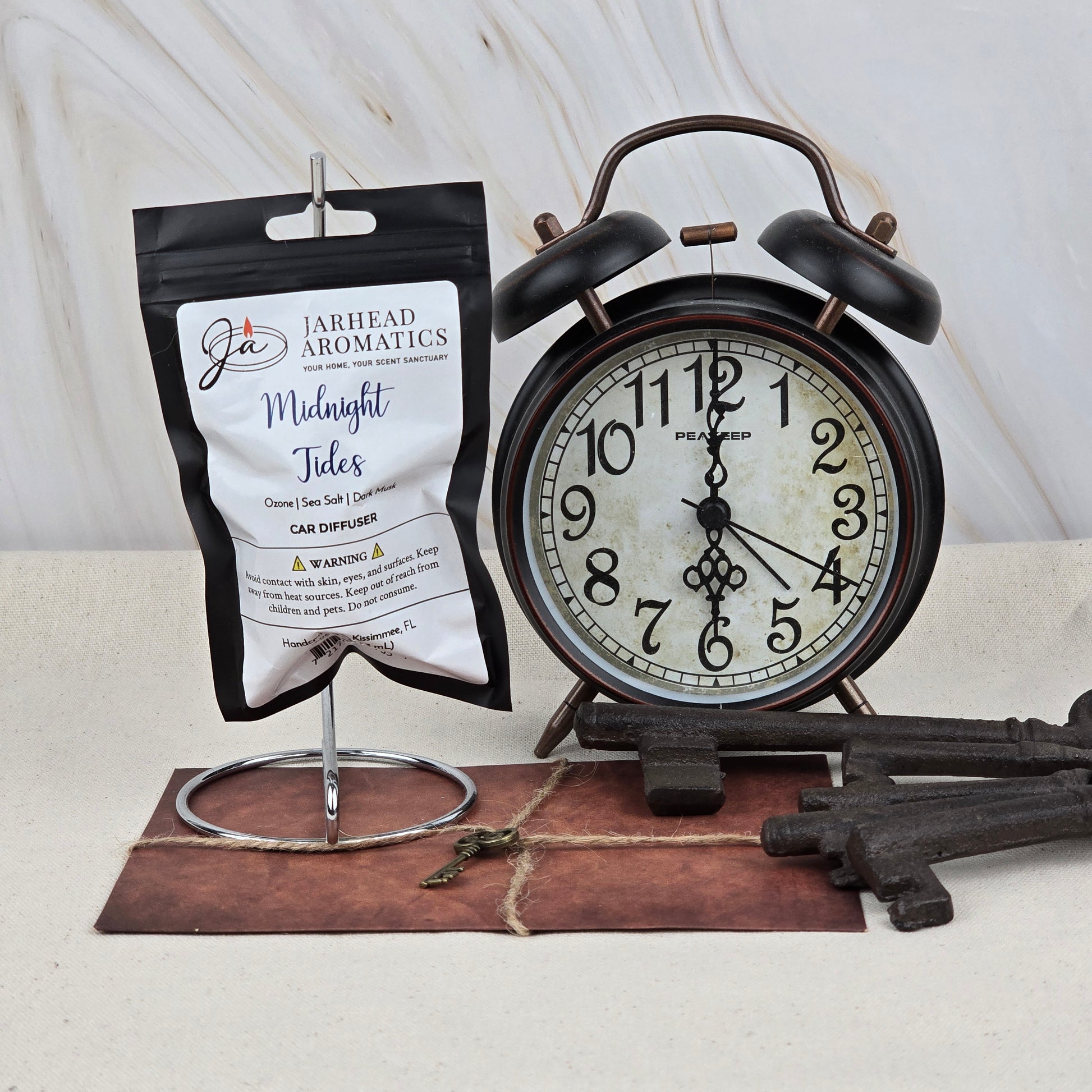 A black alarm clock displays 10:12 beside a Jarhead Aromatics Midnight Tides Car Diffuser—an Ocean Mist, Dark Musk & Sandalwood car air freshener. Large vintage keys and a small key rest on brown envelopes on a light surface.