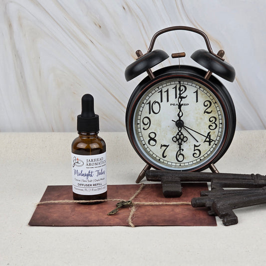 A vintage alarm clock, three large antique keys, an old envelope with a small key, and a bottle of Jarhead Aromatics Midnight Tides Car Diffuser Refill (Ocean Musk & Sandalwood) are arranged on a light surface against a marble background.