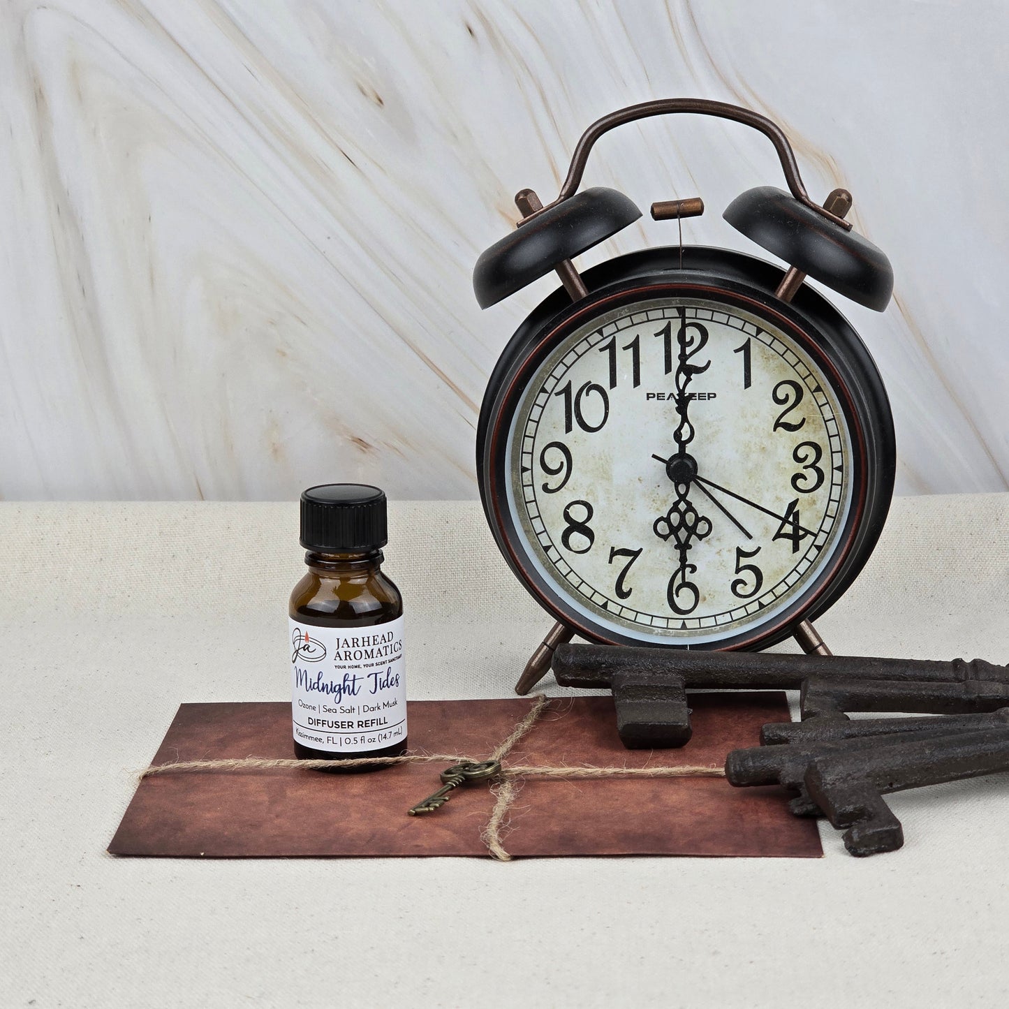 A vintage-style black alarm clock, a large old-fashioned key, an envelope with string, and a Jarhead Aromatics Midnight Tides Cold Air Diffuser Refill (Sea Salt & Sandalwood) are arranged on a light surface with a marble-patterned background.