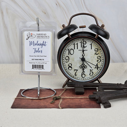A vintage black alarm clock shows 7:58 beside a Jarhead Aromatics Midnight Tides Soy Wax Melt pack—Dark Ocean Musk & Sandalwood scent—plus an old metal key and a string-tied brown envelope on a light surface.