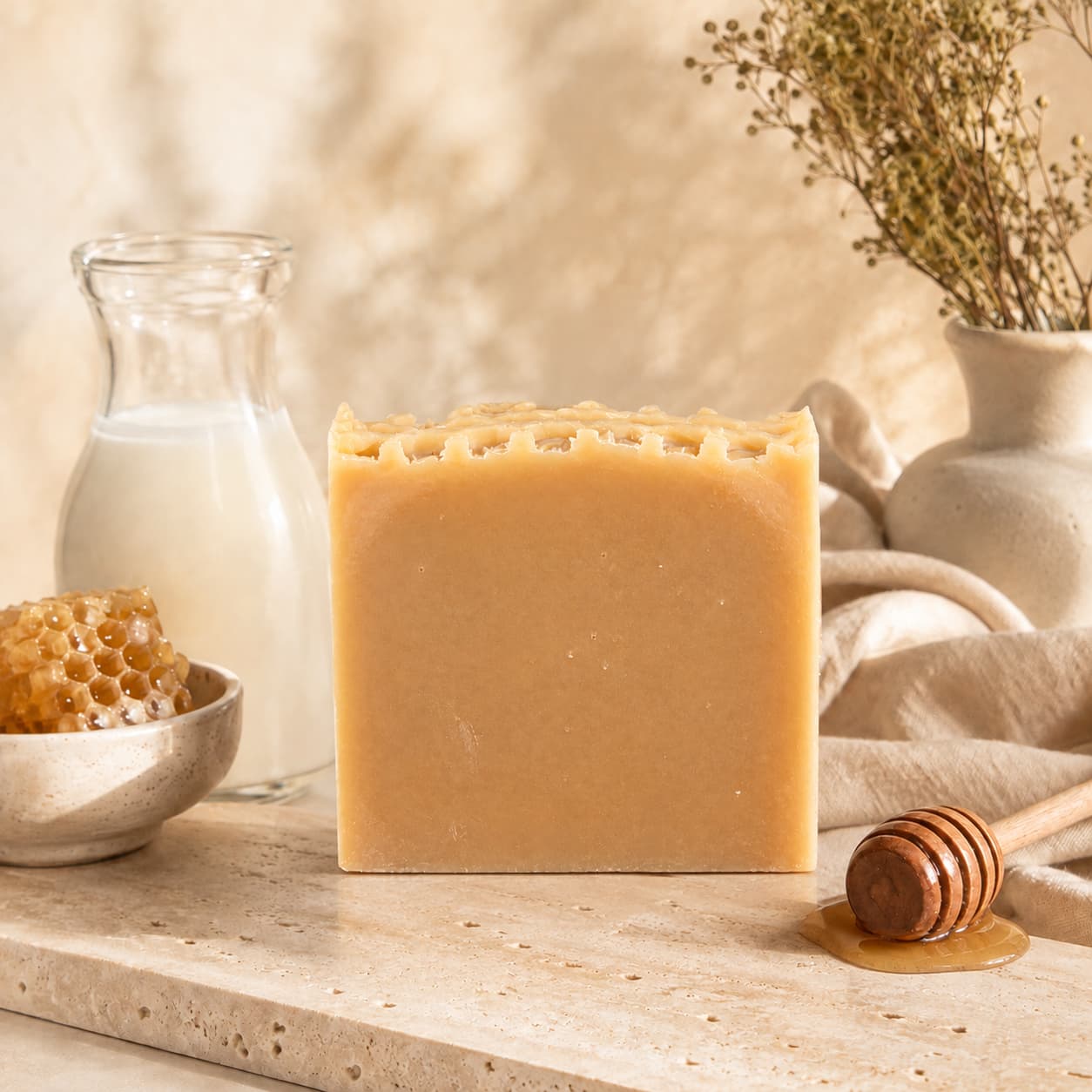 A bar of Jarhead Aromatics Milk + Honey Handmade Natural Goat Milk Artisan Soap rests on a stone surface, with a glass jug of milk, honeycomb in a bowl, a wooden honey dipper, and dried flowers arranged in the background.
