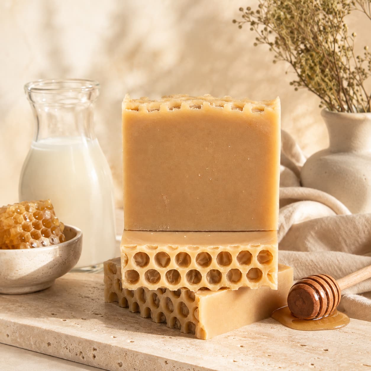 Three Milk + Honey Handmade Natural Goat Milk Artisan Soap bars by Jarhead Aromatics, featuring a honeycomb texture, are stacked on a stone surface alongside a honey dipper, a bowl of honeycomb, a small glass pitcher of milk, and a vase with dried flowers in the background.