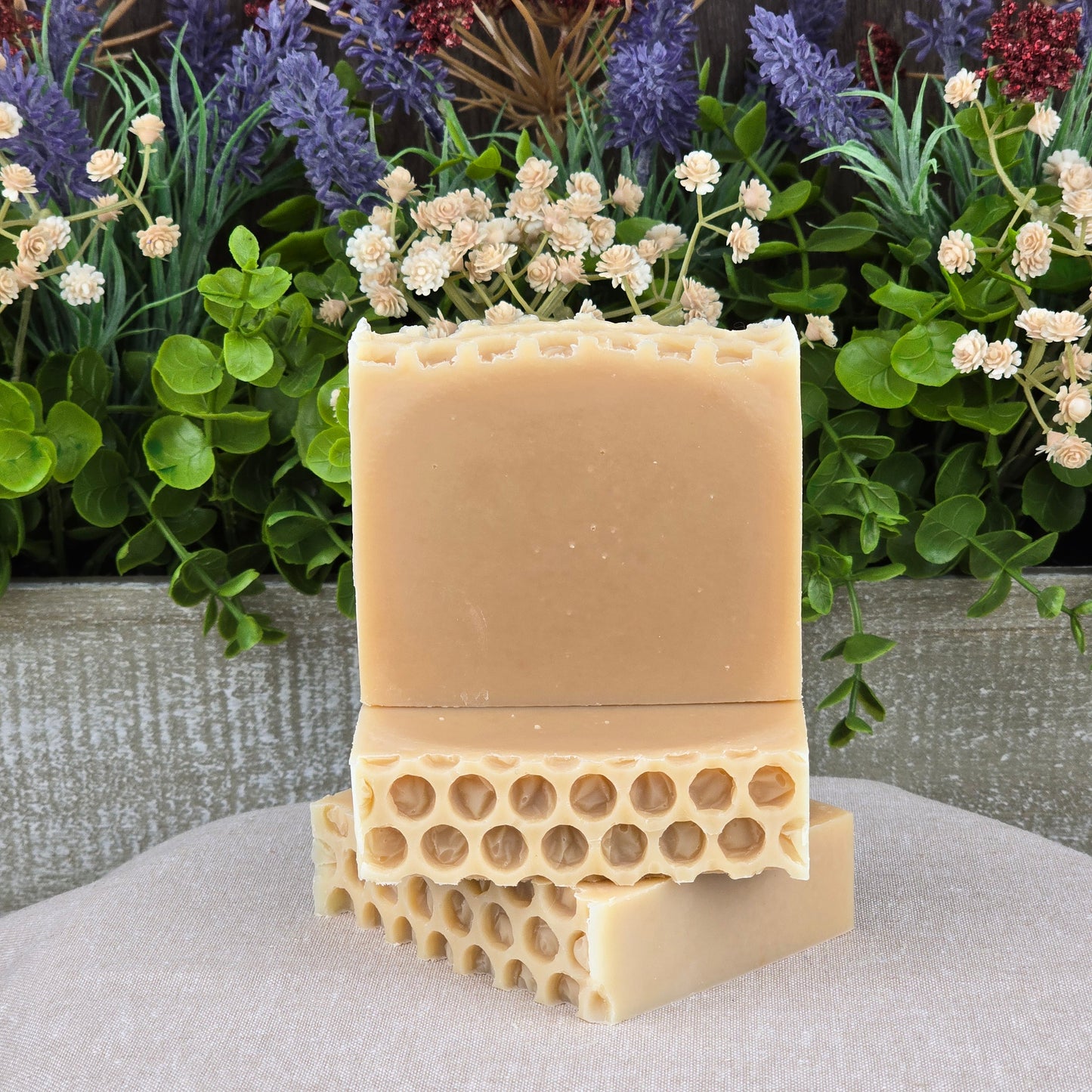 Two Milk + Honey Handmade Natural Goat Milk Artisan Soaps by Jarhead Aromatics, featuring a honeycomb pattern, are stacked on light gray fabric with green leaves and purple-white artificial flowers in the background.