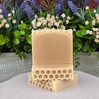 Two Milk + Honey Handmade Natural Goat Milk Artisan Soaps by Jarhead Aromatics, featuring a honeycomb pattern, are stacked on light gray fabric with green leaves and purple-white artificial flowers in the background.