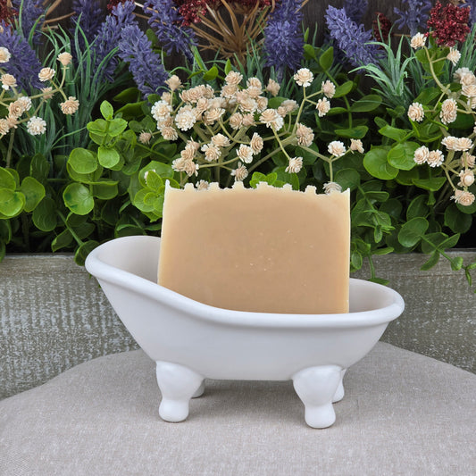 A bar of Jarhead Aromatics Milk + Honey Handmade Natural Goat Milk Artisan Soap sits in a white clawfoot bathtub-shaped dish, with lush green leaves and purple and cream artificial flowers in the background.