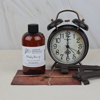 A bottle of Jarhead Aromatics Minty Breeze Diffuser Refill | Peppermint, Eucalyptus & Vanilla Home Fragrance Oil sits by a vintage black alarm clock, old-fashioned keys, and a closed leather-bound journal, creating a refreshing, spa-like ambiance.