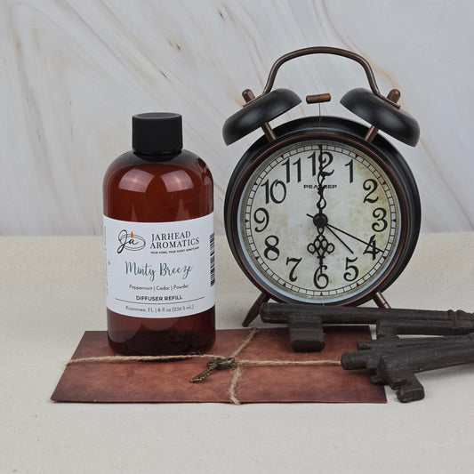 A bottle of Jarhead Aromatics Minty Breeze Diffuser Refill | Peppermint, Eucalyptus & Vanilla Home Fragrance Oil sits by a vintage black alarm clock, old-fashioned keys, and a closed leather-bound journal, creating a refreshing, spa-like ambiance.
