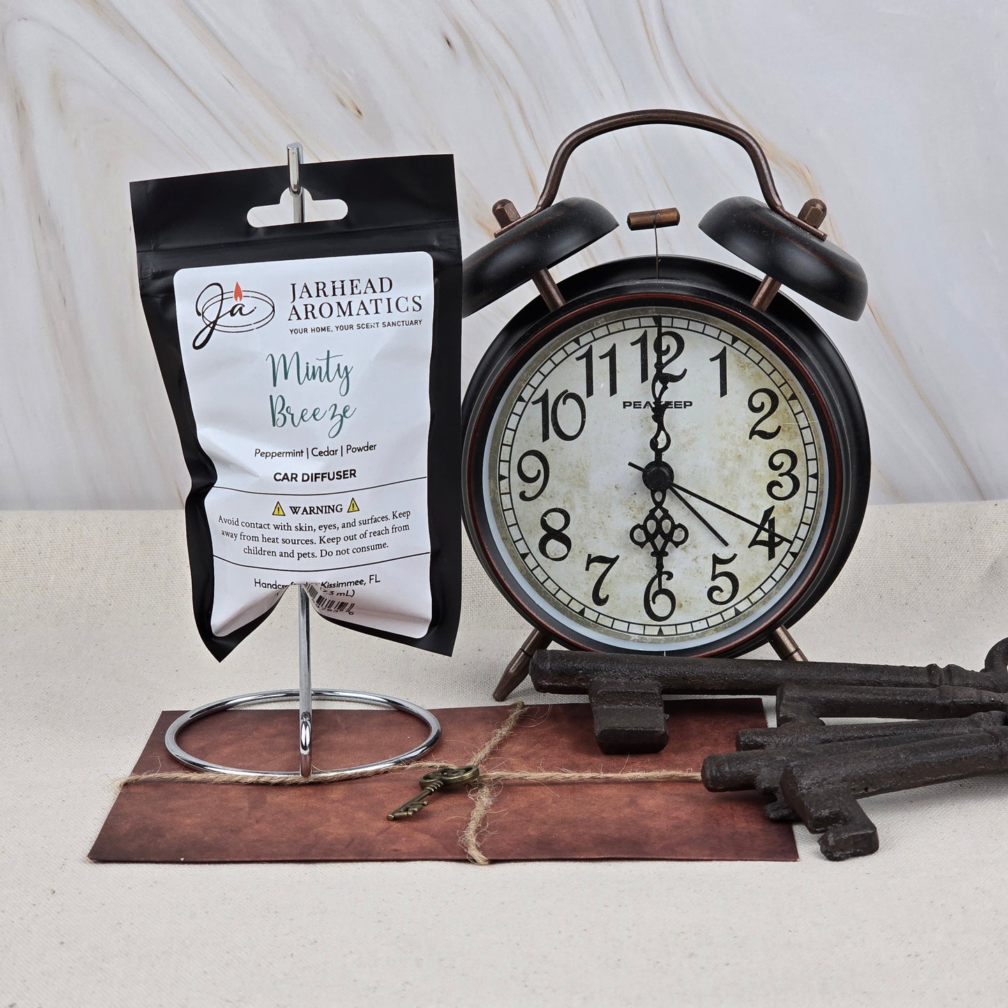 A black vintage alarm clock sits beside a Jarhead Aromatics Minty Breeze Car Diffuser—Peppermint & Eucalyptus Car Air Freshener—displayed with large old-fashioned keys and a brown envelope on a light marbled surface.
