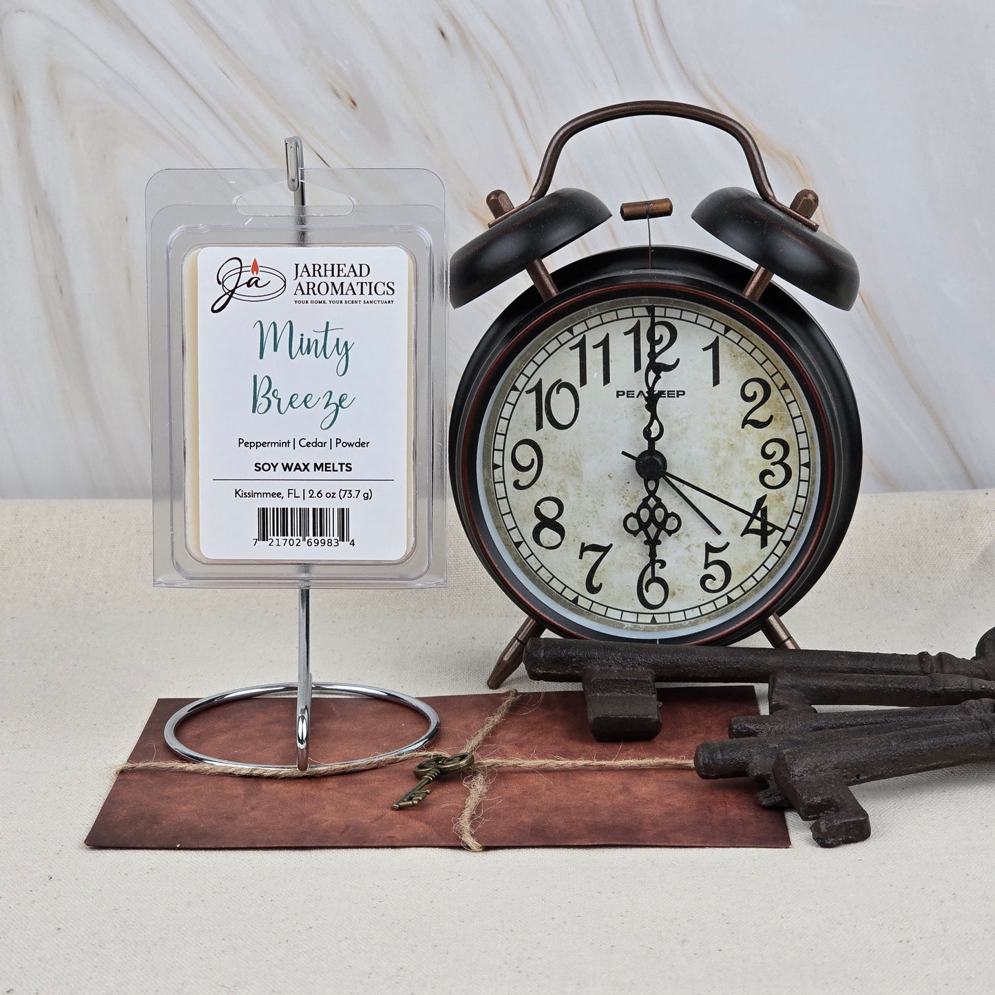 A pack of Jarhead Aromatics Minty Breeze Soy Wax Melt | Peppermint Eucalyptus Spa Wax Melts rests on a holder by a vintage black alarm clock and decorative keys, bringing long-lasting fragrance on a brown surface with a marble backdrop.