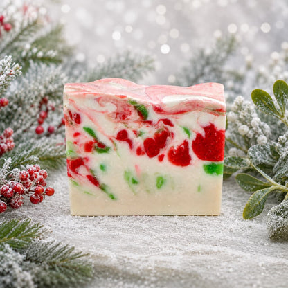 A bar of swirled Mistletoe Handmade Artisan Soap in red, white, and green sits on a snowy surface surrounded by pine branches and holly berries.