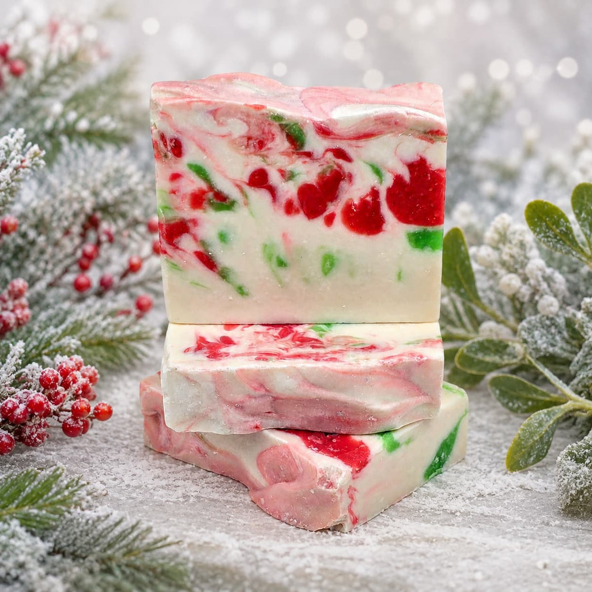 Three rectangular bars of Mistletoe Handmade Artisan Soap with red and green swirls, stacked on a snowy surface decorated with holly and mistletoe.