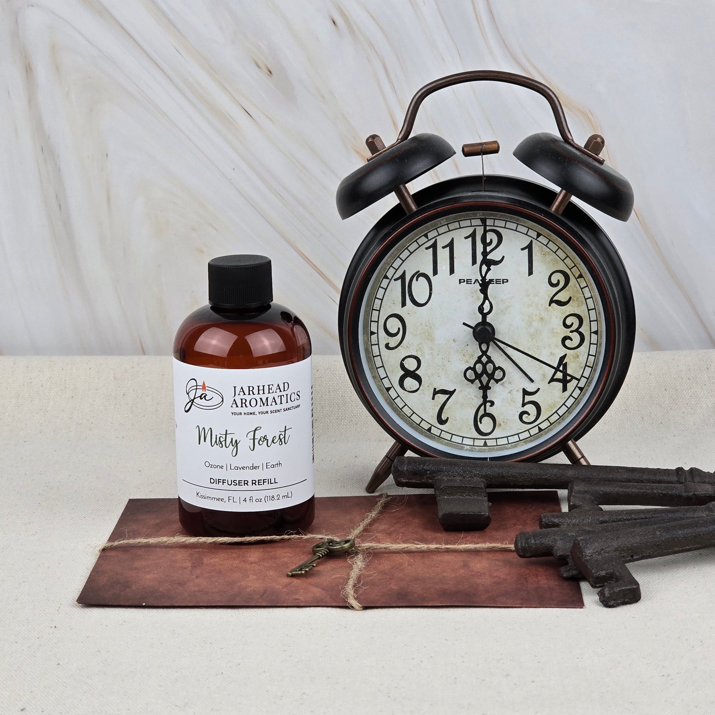 A Jarhead Aromatics Misty Forest Diffuser Refill—Ozone, Lavender & Juniper Home Fragrance Oil—sits alongside an alarm clock, vintage keys, and a leather-bound book on a light surface with a marble background, evoking woodland aromas.