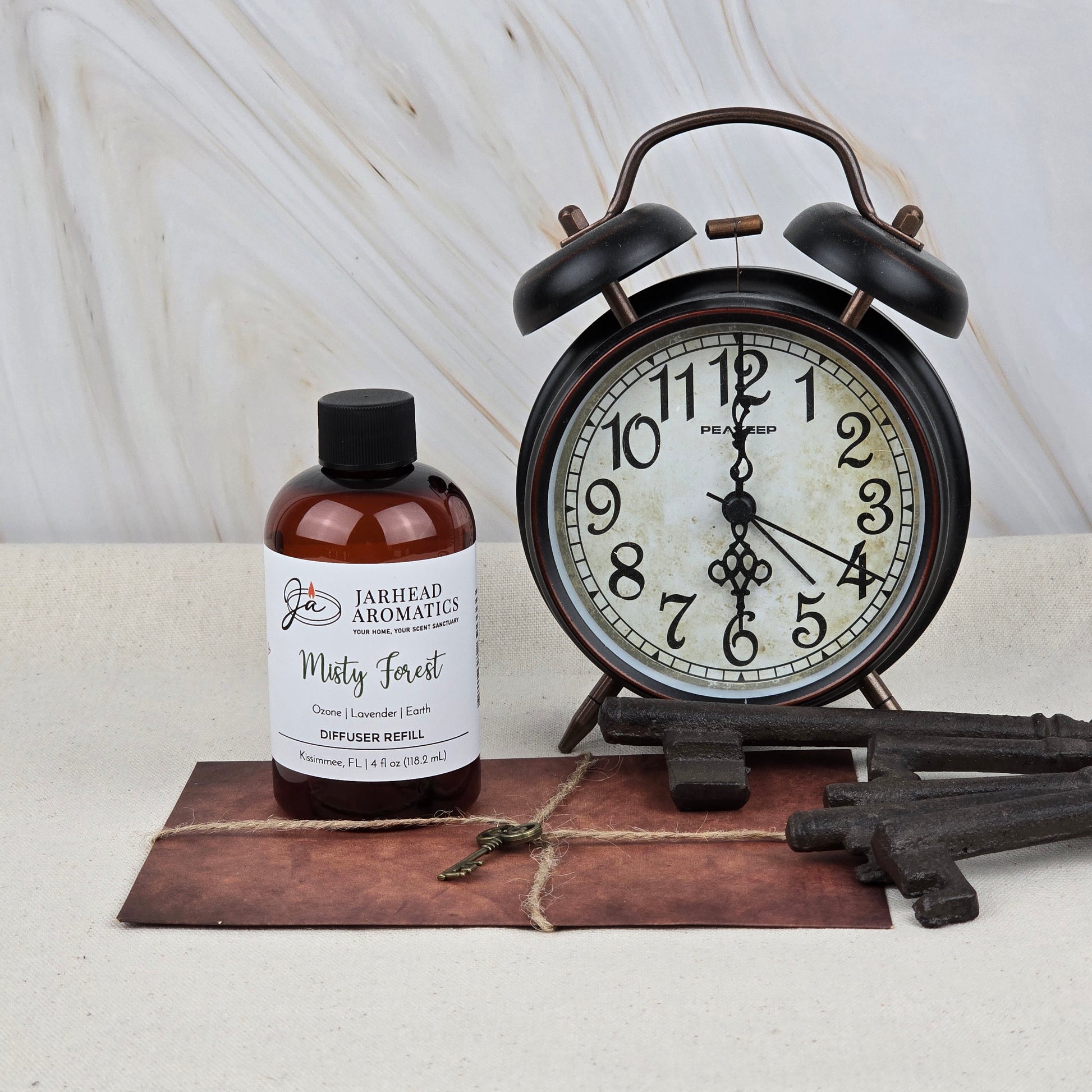A Jarhead Aromatics Misty Forest Diffuser Refill—Ozone, Lavender & Juniper Home Fragrance Oil—sits alongside an alarm clock, vintage keys, and a leather-bound book on a light surface with a marble background, evoking woodland aromas.