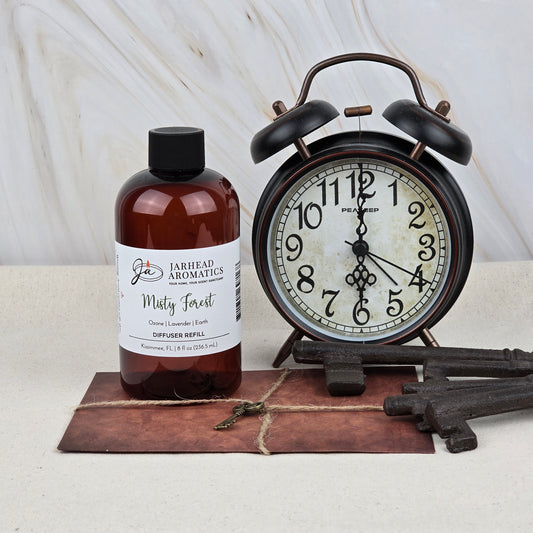 A brown bottle of Jarhead Aromatics Misty Forest Diffuser Refill | Ozone, Lavender & Juniper sits beside a black vintage alarm clock, old metal keys, and a leather-bound book, evoking the earthy aroma of a woodland retreat.