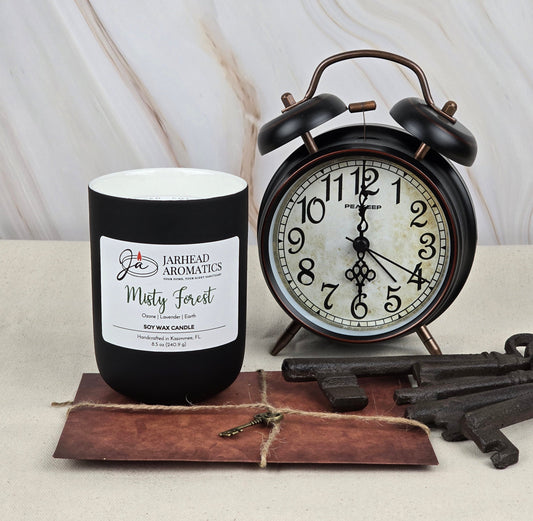 A Misty Forest soy wax candle by Jarhead Aromatics emits a refreshing aroma with essential oils, perched next to a vintage-style black alarm clock with a white face and rustic metal keys on vintage paper.
