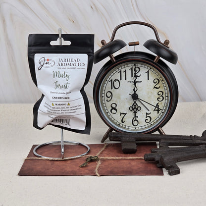 A black vintage alarm clock shows 2:55 beside a Jarhead Aromatics Misty Forest Car Diffuser—an earthy woodland, rain-kissed air freshener—hanging on a stand. Large decorative keys and a small antique key rest on a leather-bound pad below.