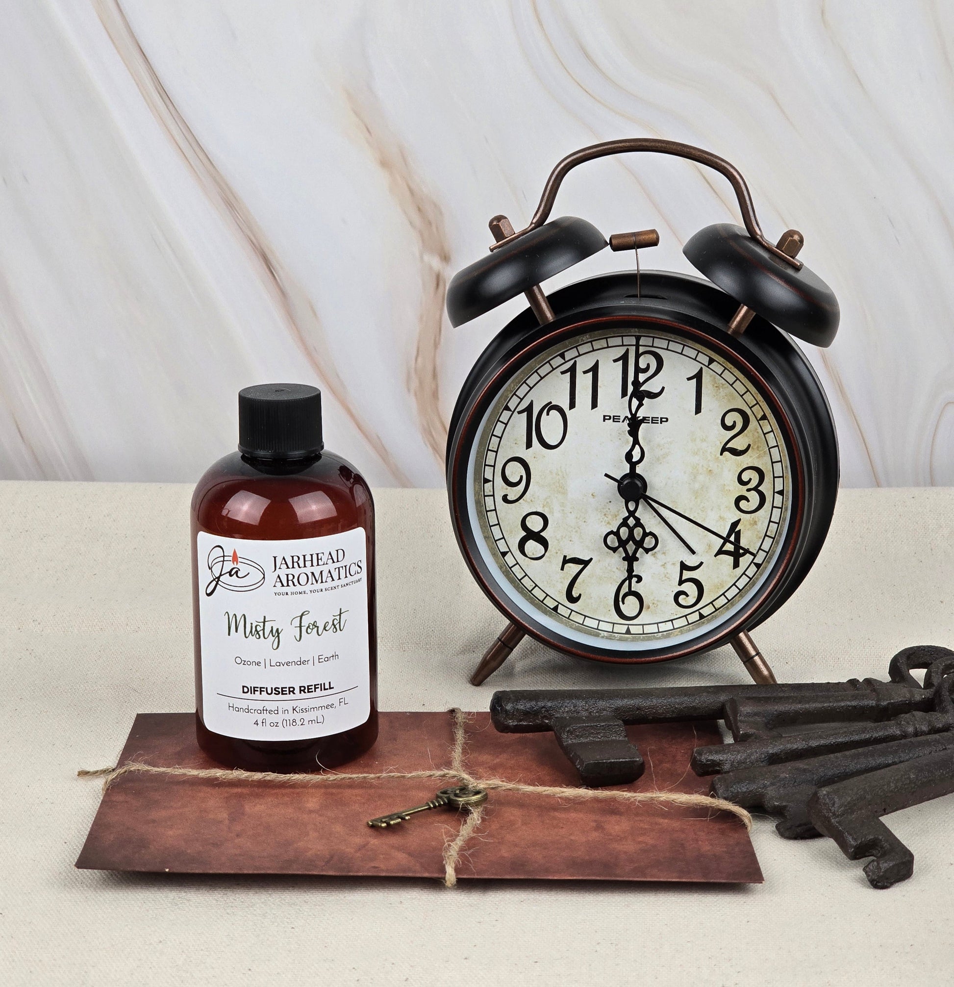 A vintage black clock showing 11:04 sits next to a Misty Forest - Reed Diffuser Refill by Jarhead Aromatics, emanating an earthy scent. The bottle lies on a brown leather-like mat with three skeleton keys, against a light marble backdrop.