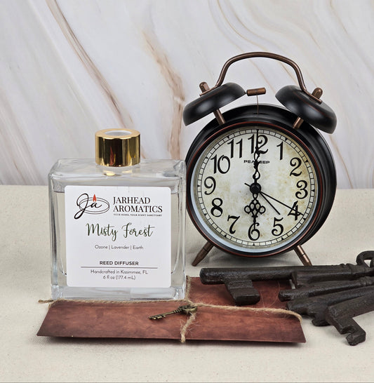 A Jarhead Aromatics Misty Forest reed diffuser, with its earthy essential oils, rests on a brown envelope beside a vintage black alarm clock and metal keys on a beige marbled surface, highlighting its natural charm.