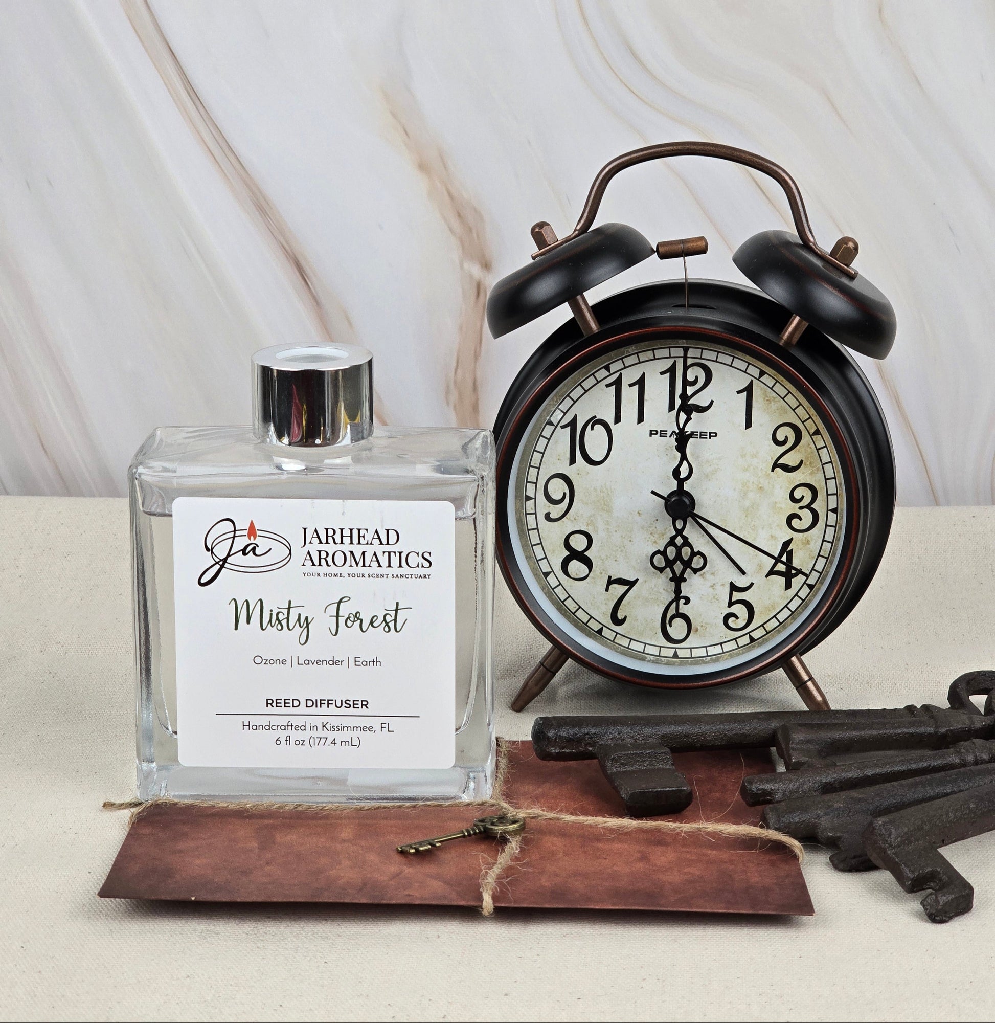 A Jarhead Aromatics Misty Forest - Reed Diffuser with earthy essential oils is displayed next to an antique black alarm clock and vintage keys on a white surface. A brown card sits beneath the diffuser against a marbled backdrop.