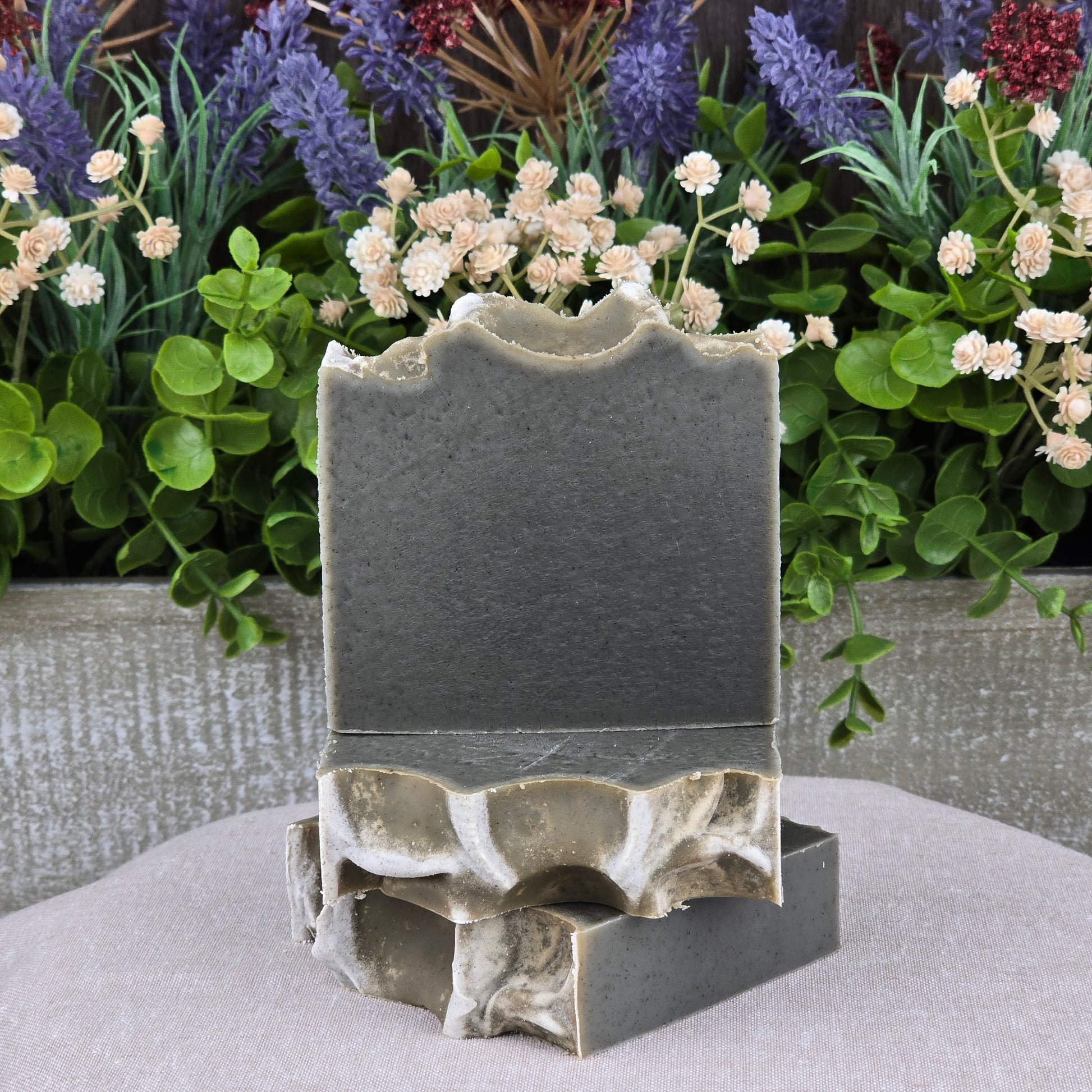 Two Mugwort + Lavender Handmade Natural Artisan Soaps by Jarhead Aromatics with wavy tops are stacked before lush greenery and blooming purple and white flowers, highlighting their artisanal elegance and natural ingredients.