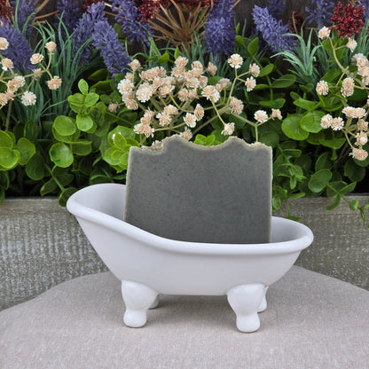 A gray bar of Jarhead Aromatics Mugwort + Lavender Handmade Natural Artisan Soap rests in a small white bathtub-shaped soap dish, with green and purple faux plants and flowers in the background.