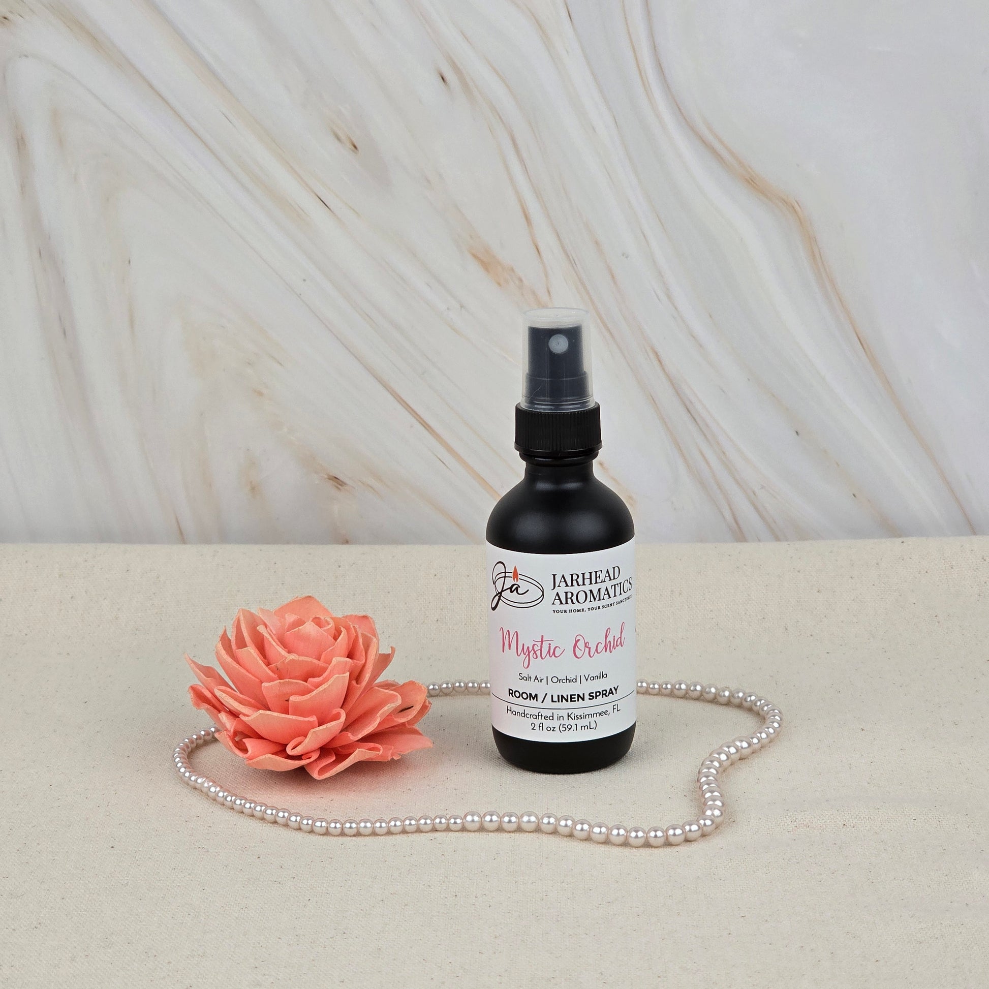 A bottle of Jarhead Aromatics Mystic Orchid Room & Linen Spray (Orchid, Sea Air & Vanilla) sits on a beige surface with a pink artificial flower and pearls, evoking a fresh coastal home scent against a light marble background.
