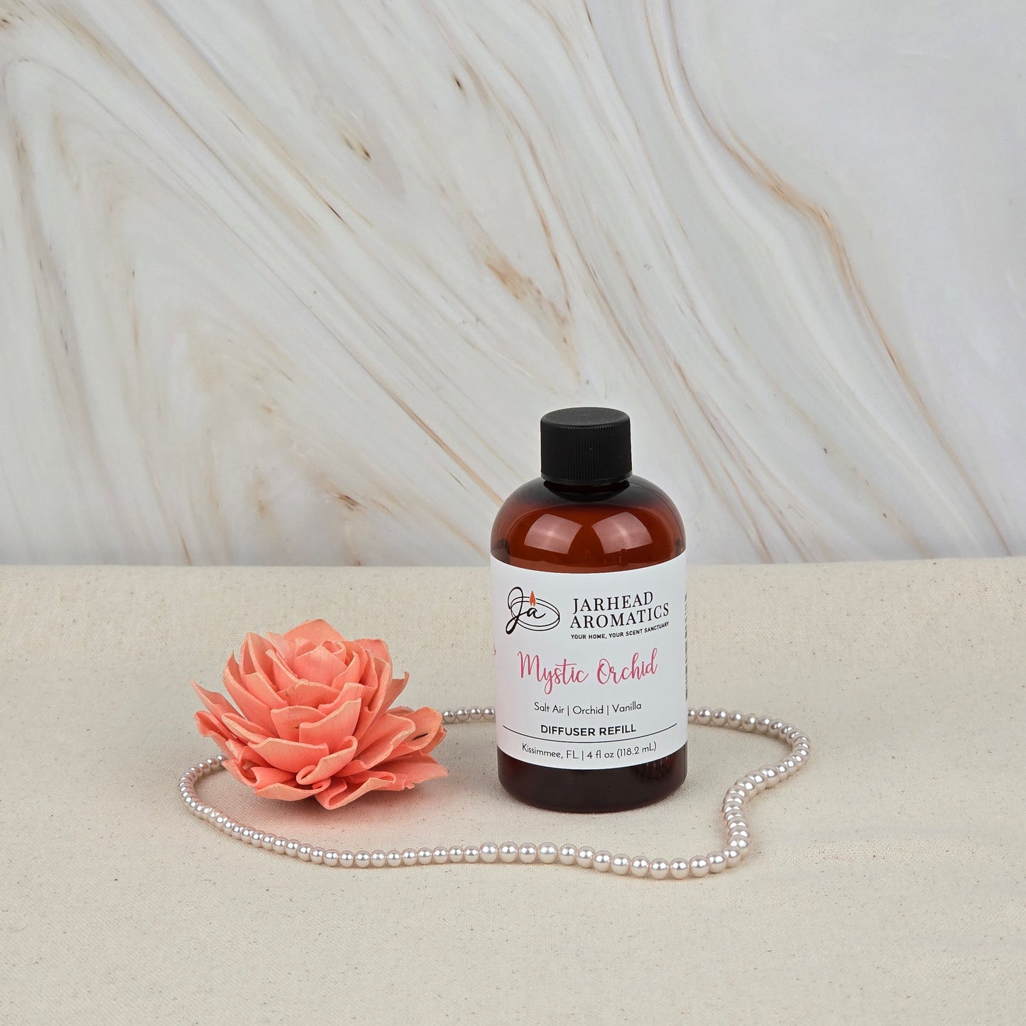 A bottle of Jarhead Aromatics Mystic Orchid Diffuser Refill—an Orchid, Sea Air & Vanilla home fragrance oil—rests on beige beside a pink artificial flower and pearls, with a marbled background reflecting coastal elegance.