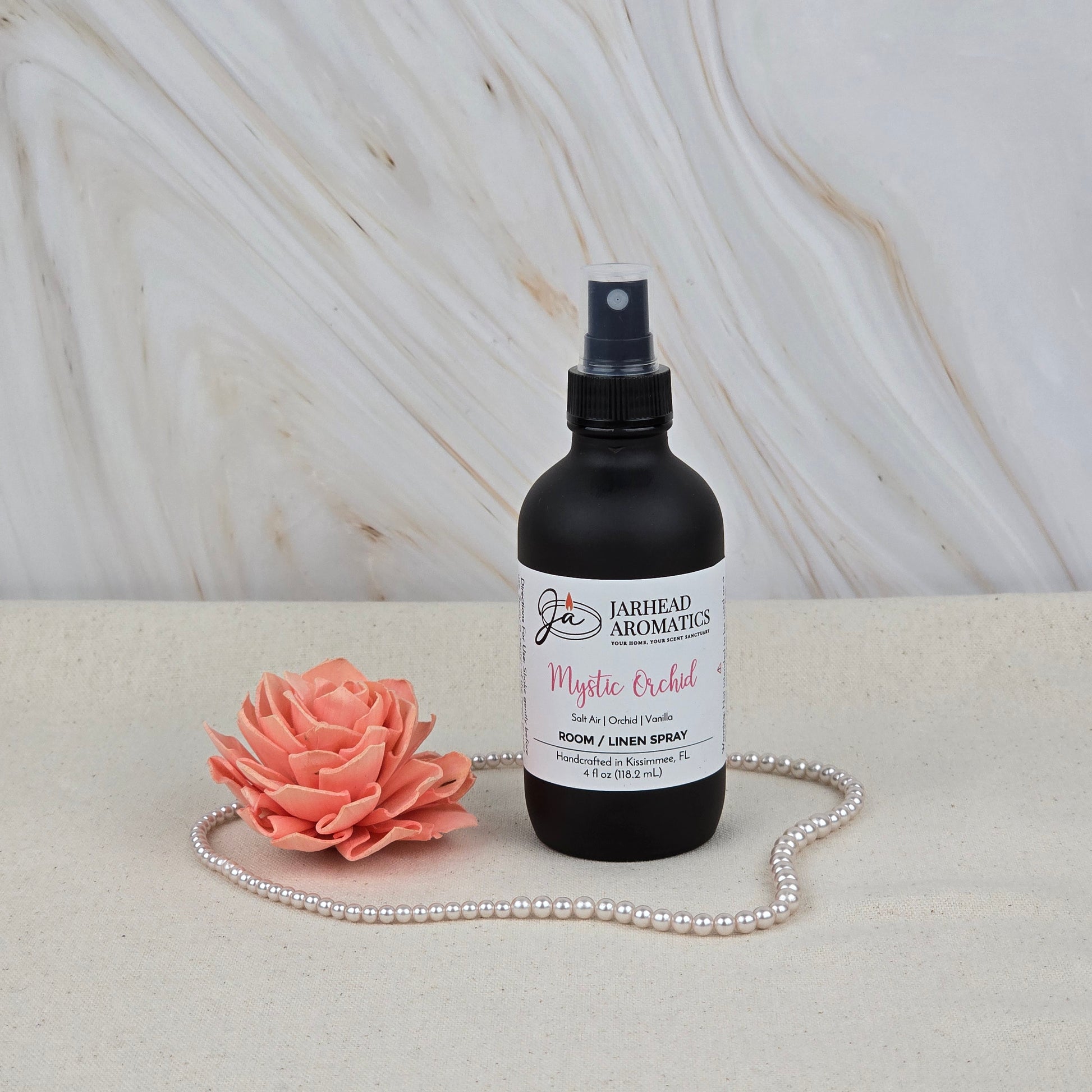 A black Jarhead Aromatics Mystic Orchid Room & Linen Spray (Orchid, Sea Air & Vanilla) rests on a light surface next to a pink artificial flower and pearls, with a marble background—ideal as a coastal home or linen fragrance.