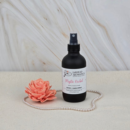A black Jarhead Aromatics Mystic Orchid Room & Linen Spray (Orchid, Sea Air & Vanilla) rests on a light surface next to a pink artificial flower and pearls, with a marble background—ideal as a coastal home or linen fragrance.