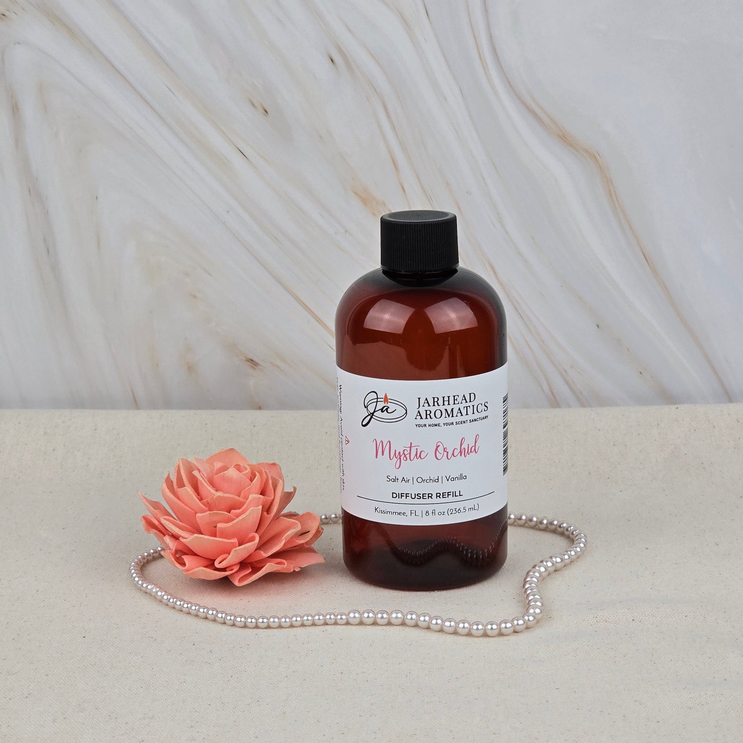 A brown bottle of Jarhead Aromatics Mystic Orchid Diffuser Refill | Orchid, Sea Air & Vanilla sits on a beige surface beside a coral flower and pearls, evoking floral elegance against the marble-patterned background.