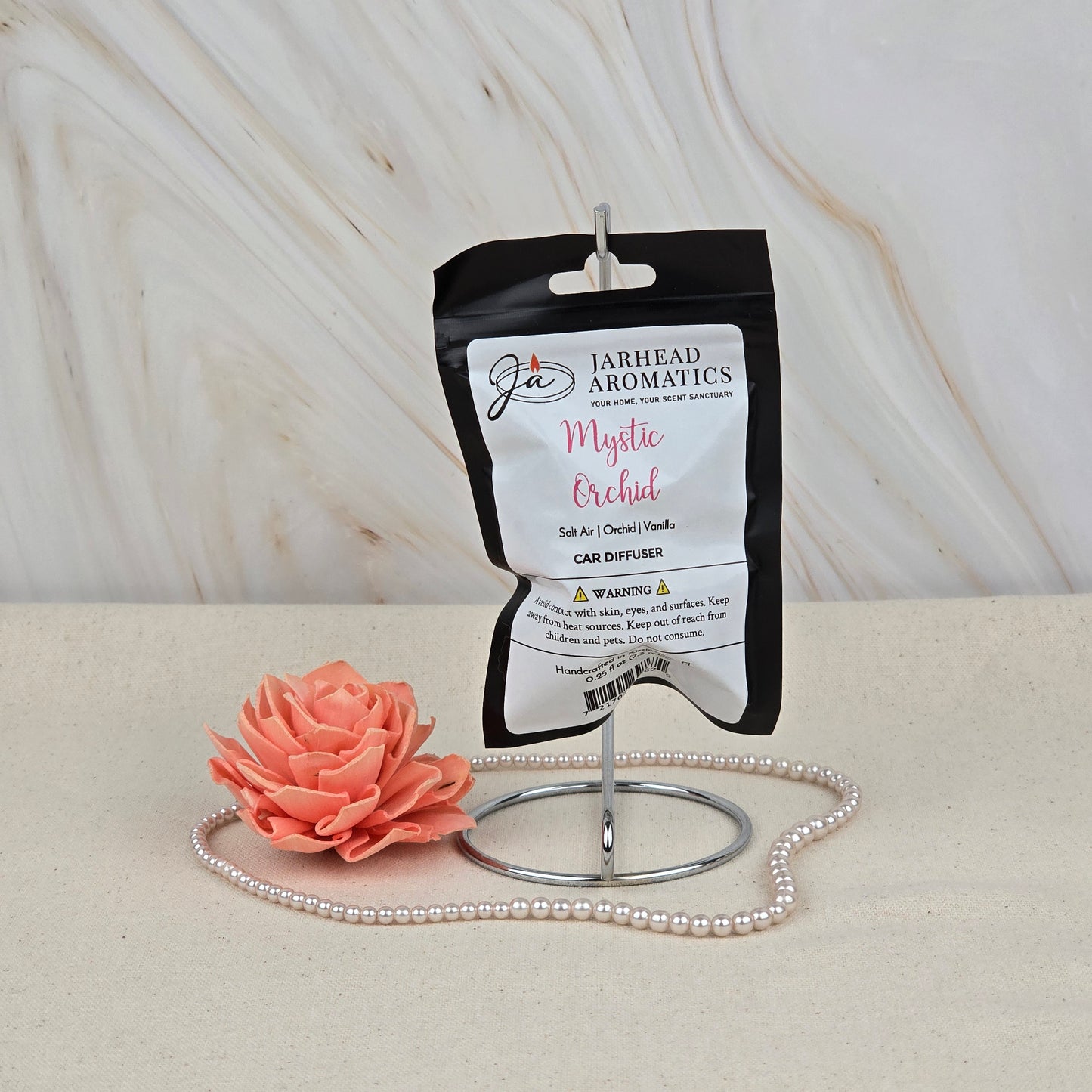 The Mystic Orchid Car Diffuser | Coastal Floral & Vanilla Musk Car Air Freshener by Jarhead Aromatics is displayed on a metal stand with a pink flower and pearls, highlighting this long-lasting car fragrance against a marbled background.