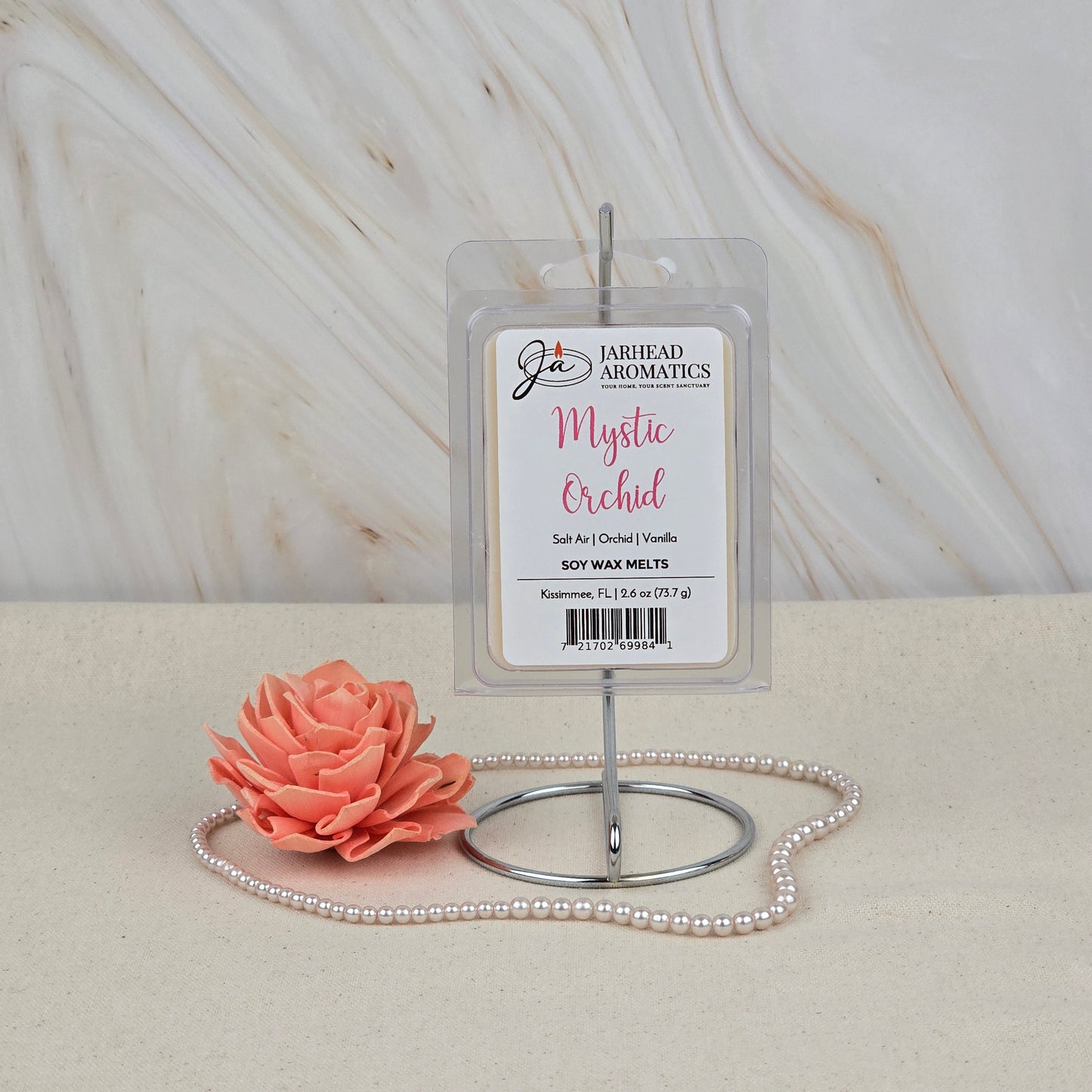 Jarhead Aromatics Mystic Orchid Soy Wax Melt | Coastal Orchid & Vanilla Wax Melts is shown on a metal stand with a pink artificial flower and pearl necklace on beige, set against marble, suggesting a fresh coastal scent.