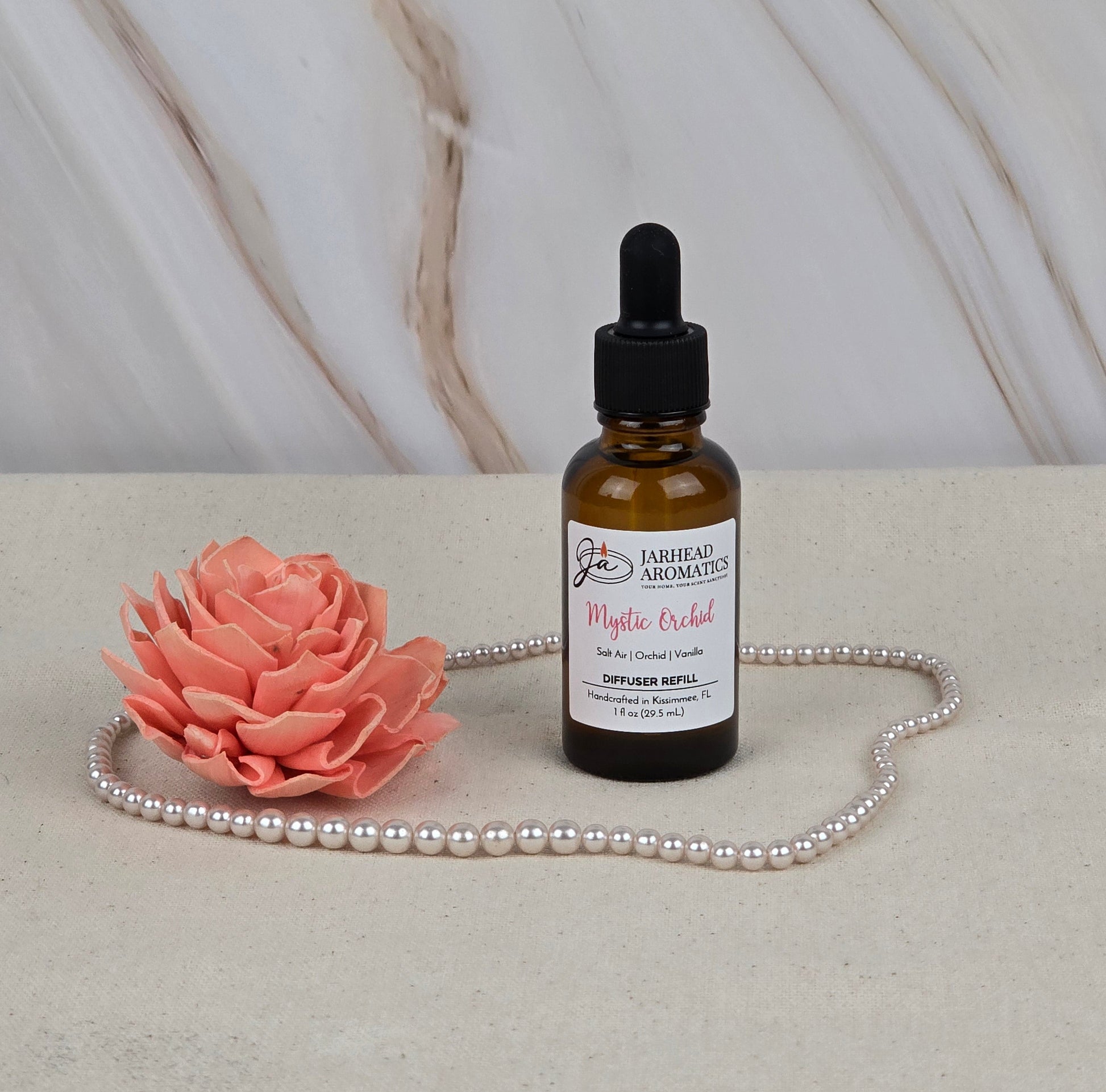 A bottle of Jarhead Aromatics Mystic Orchid Car Diffuser Refill with a dropper sits on marble, surrounded by pearls and a coral flower, blending floral mystery with coastal freshness against the sophisticated stone texture.